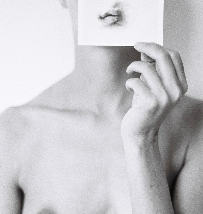 Monochrome portrait of a person holding a photo of a mouth in front of their own face, in a simple light-grey indoor setting.