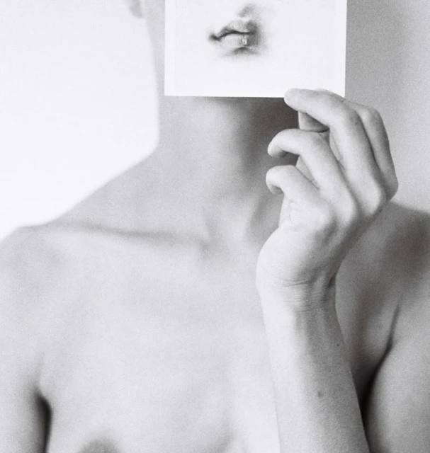 Monochrome portrait of a person holding a photo of a mouth in front of their own face, in a simple light-grey indoor setting.