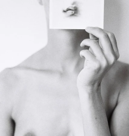 Monochrome portrait of a person holding a photo of a mouth in front of their own face, in a simple light-grey indoor setting.