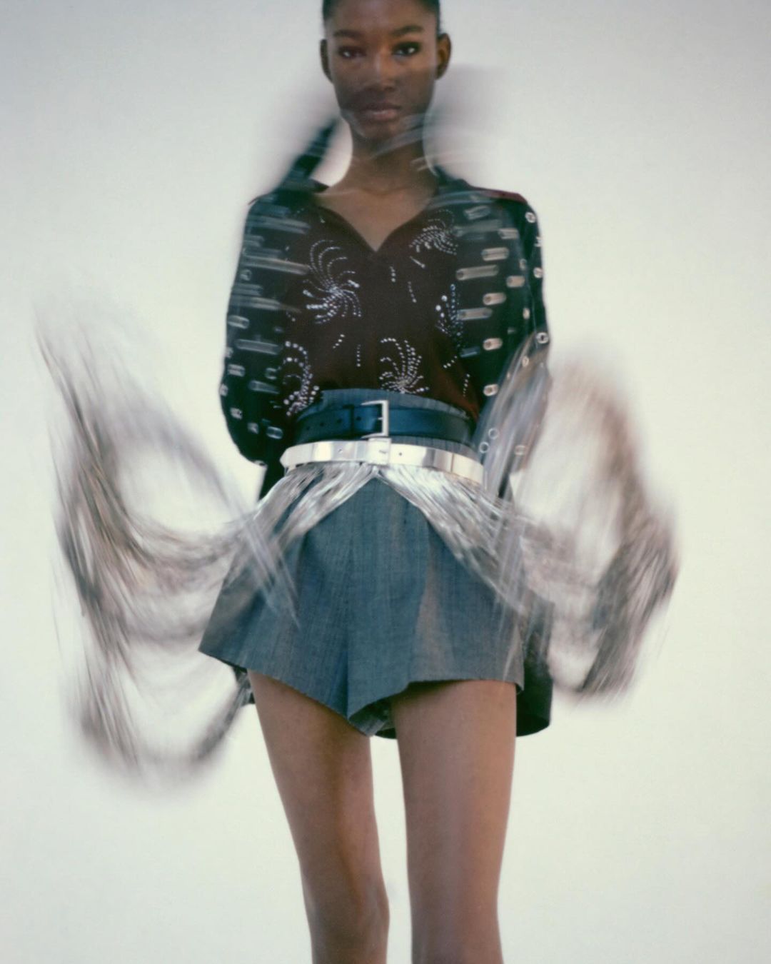 Fashion model wearing denim shorts and embellished top posing in a studio with blurred motion, against a clean white backdrop.