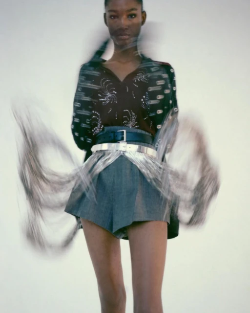 Fashion model wearing denim shorts and embellished top posing in a studio with blurred motion, against a clean white backdrop.