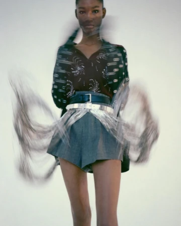 Fashion model wearing denim shorts and embellished top posing in a studio with blurred motion, against a clean white backdrop.