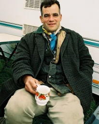 Man seated outdoors holding a patterned mug, wearing a dark green coat and light trousers with a blue neck scarf.