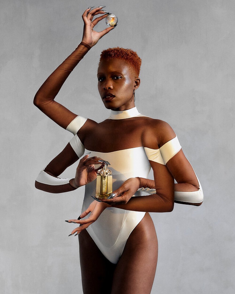 A surreal fashion photo features a person with multiple arms holding a light bulb and perfume bottle against a gray studio backdrop.