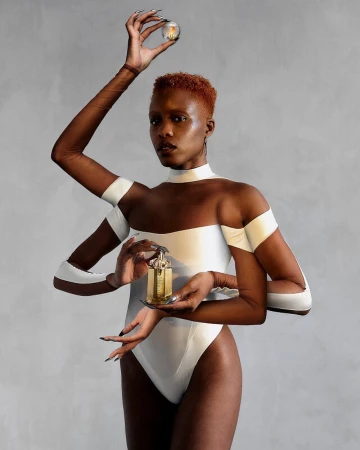 A surreal fashion photo features a person with multiple arms holding a light bulb and perfume bottle against a gray studio backdrop.
