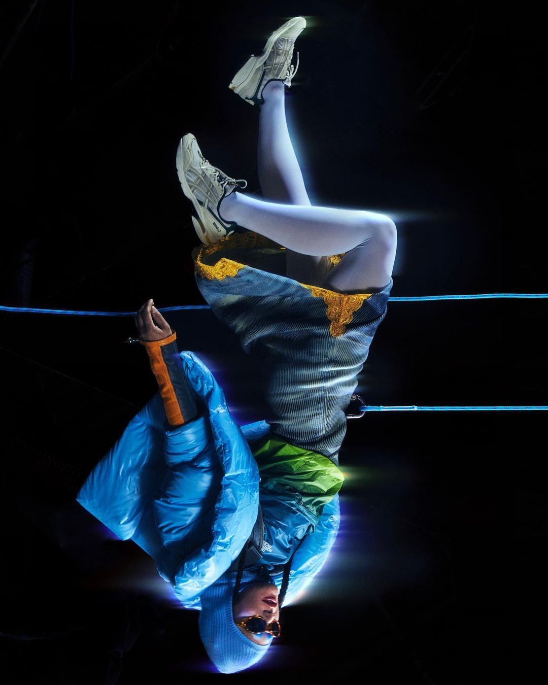 Upside-down shot of an adult wearing a blue puffer jacket, patterned skirt, and grey sneakers, with a black background.