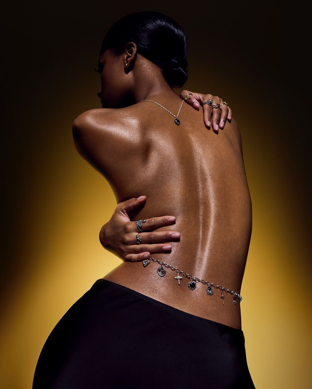 Back view of a sleek dark-skinned woman adorned with a silver necklace, rings, and body chain against a gradient yellow background.