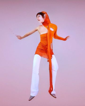 Woman in an orange top and white pants, posing in a studio. She is wearing an orange scarf-like appendage with holes and a liquid texture.