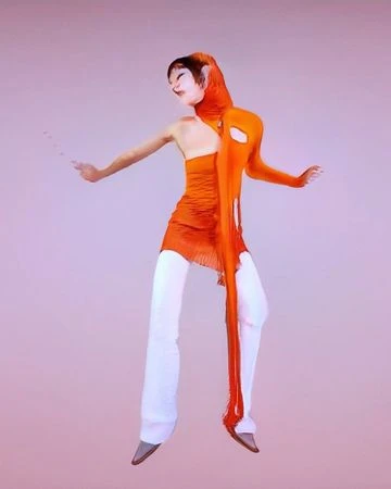 Woman in an orange top and white pants, posing in a studio. She is wearing an orange scarf-like appendage with holes and a liquid texture.