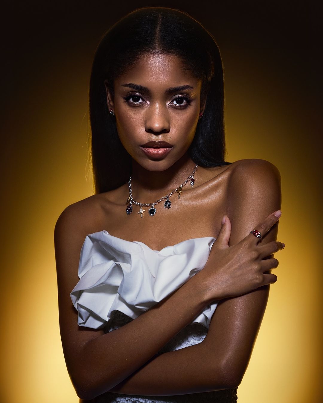 Portrait of a young woman with dark hair in an off-the-shoulder white top against a golden-yellow gradient backdrop in a studio setting.