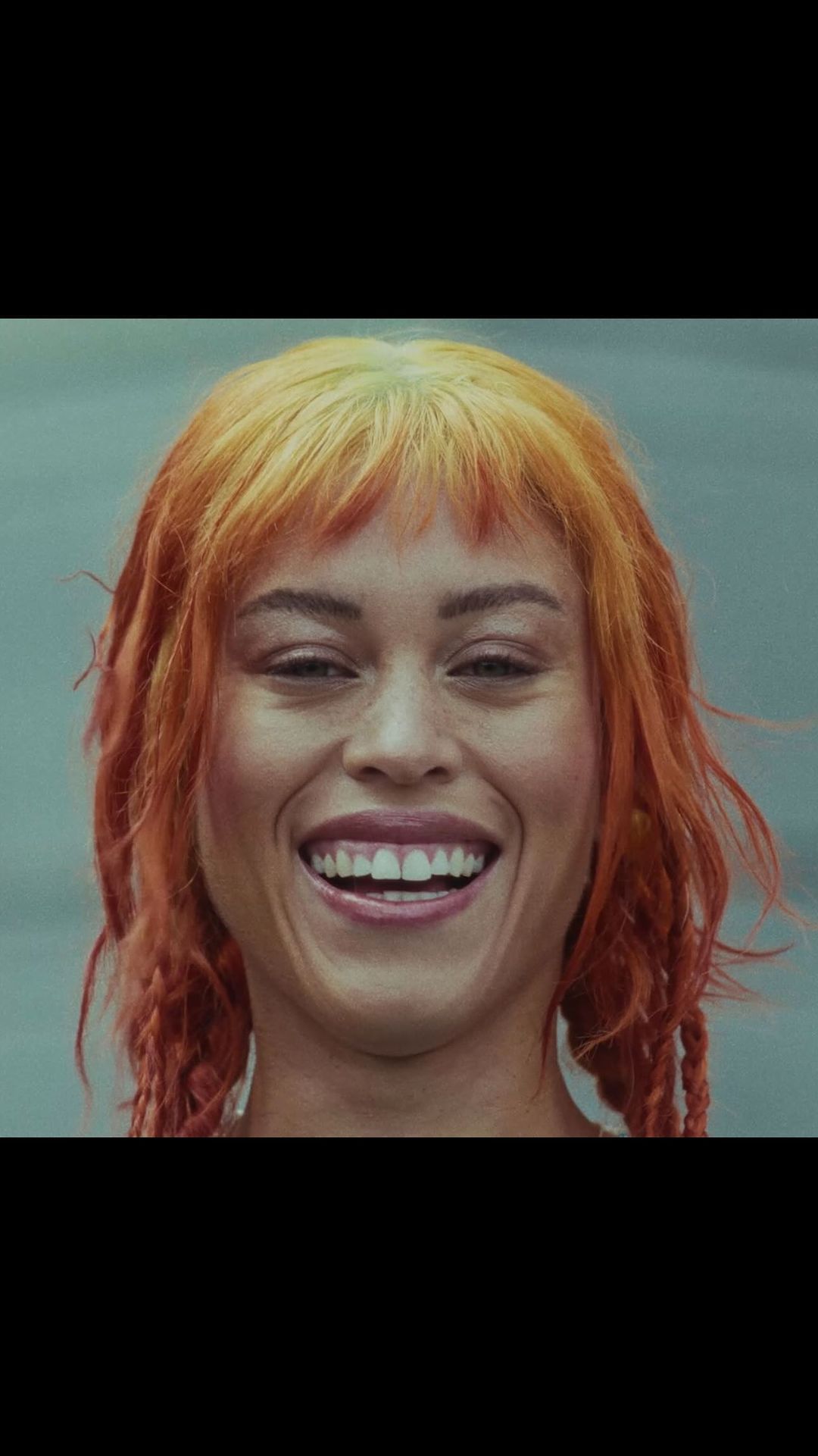 Close-up portrait of a smiling adult female with orange hair and braided pigtails against a blurred, light background.