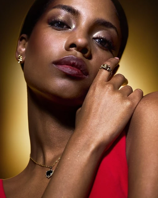 Close-up studio portrait of a Black woman with golden eye makeup, a red top, and jewelry including earrings, a necklace, and a ring.