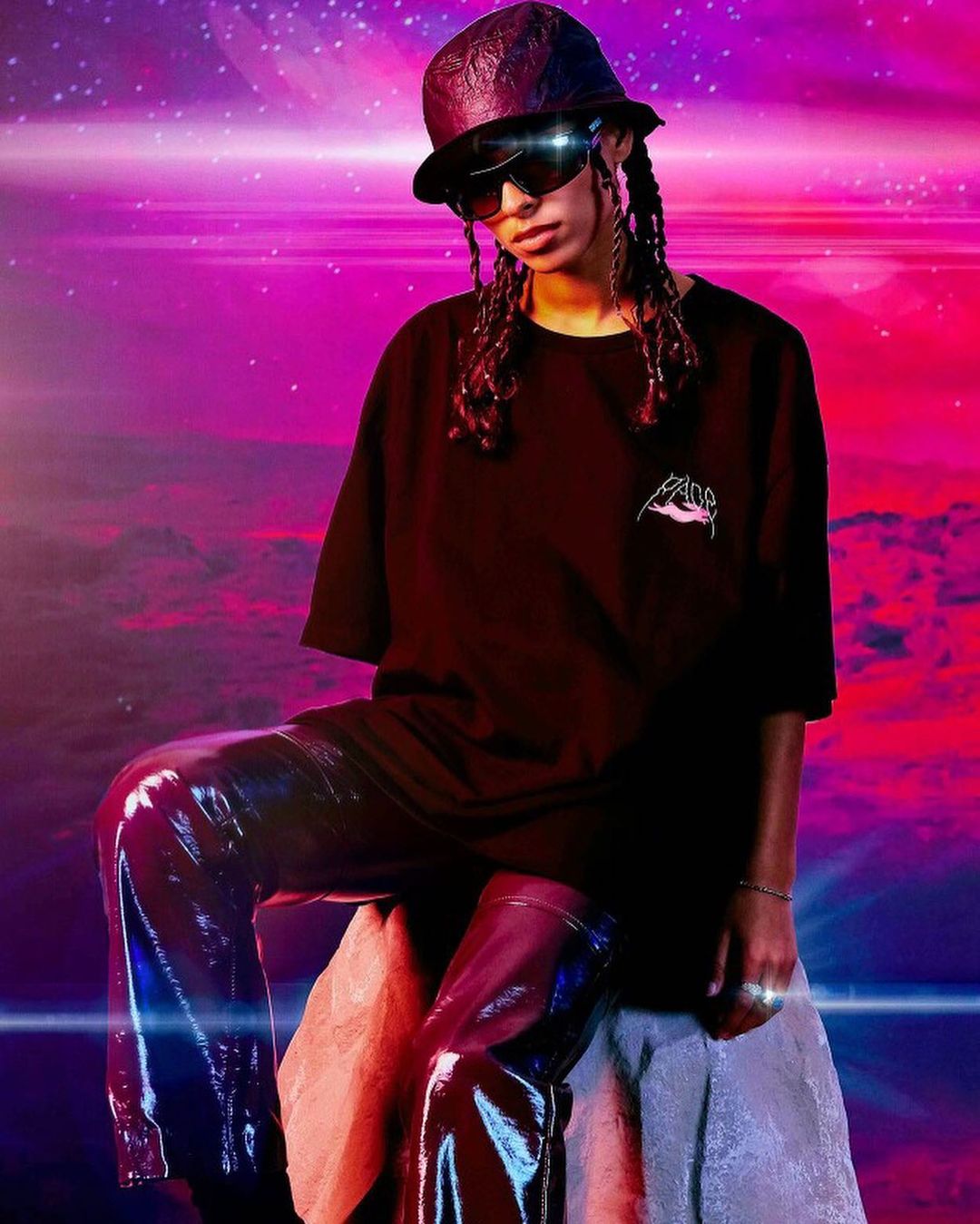 A person with locs wears sunglasses, a bucket hat, and a T-shirt with logo art, while posing against a blurred, colorful background.