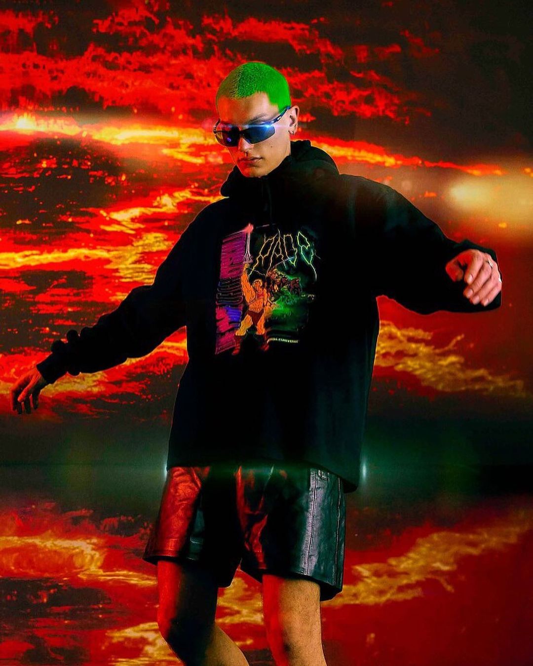 An adult male with bright green hair is posing in a black hoodie and dark shorts, against a dramatic red and orange background.
