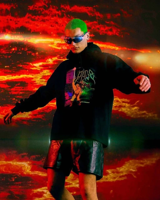 An adult male with bright green hair is posing in a black hoodie and dark shorts, against a dramatic red and orange background.
