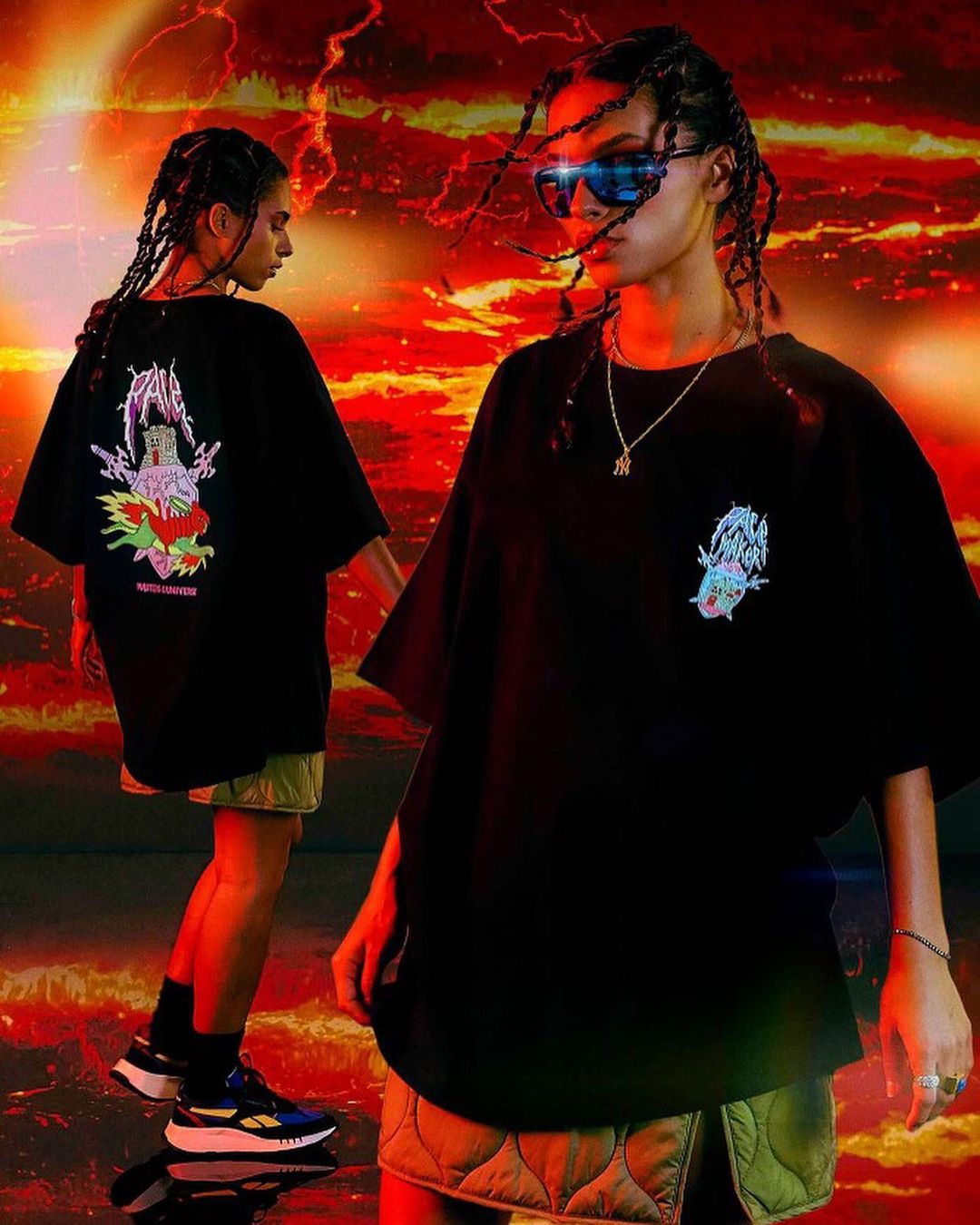 Two young adult women in black oversized T-shirts and quilted skirts pose against a fiery red and orange abstract backdrop indoors.