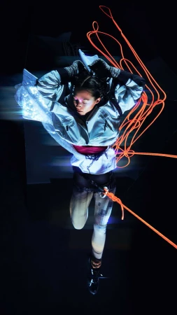 Woman poses in futuristic attire with neon rope accents in studio, creating a dynamic, high-angle full shot with dramatic lighting.
