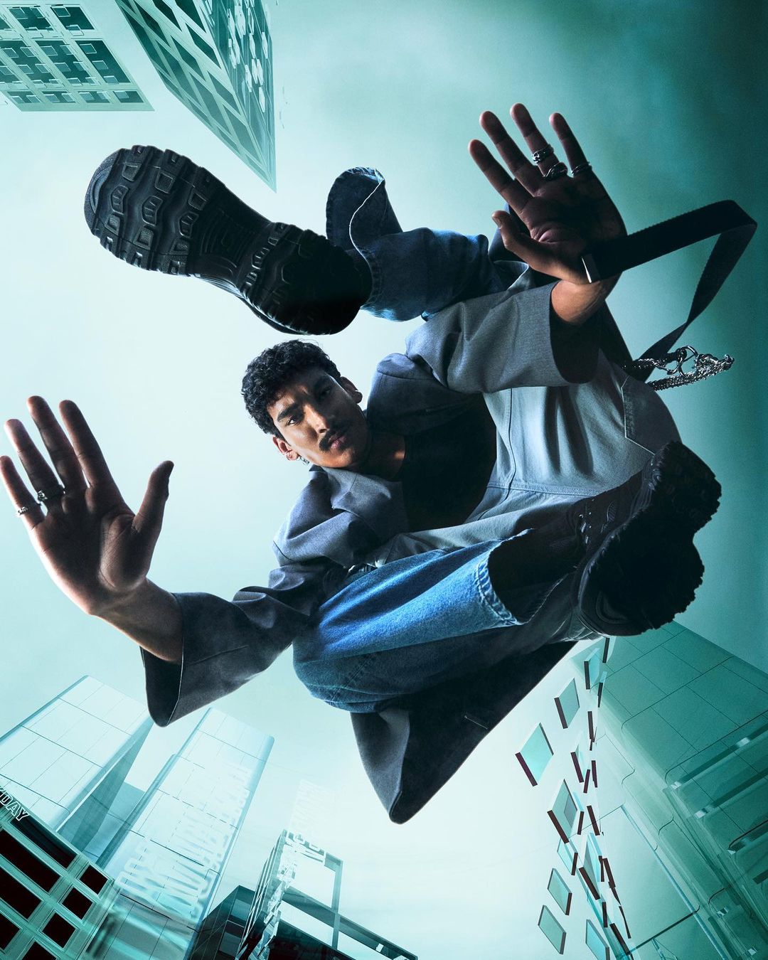 Low-angle shot of an adult man in a jacket and jeans, arms outstretched, against a backdrop of tall buildings and a teal sky.