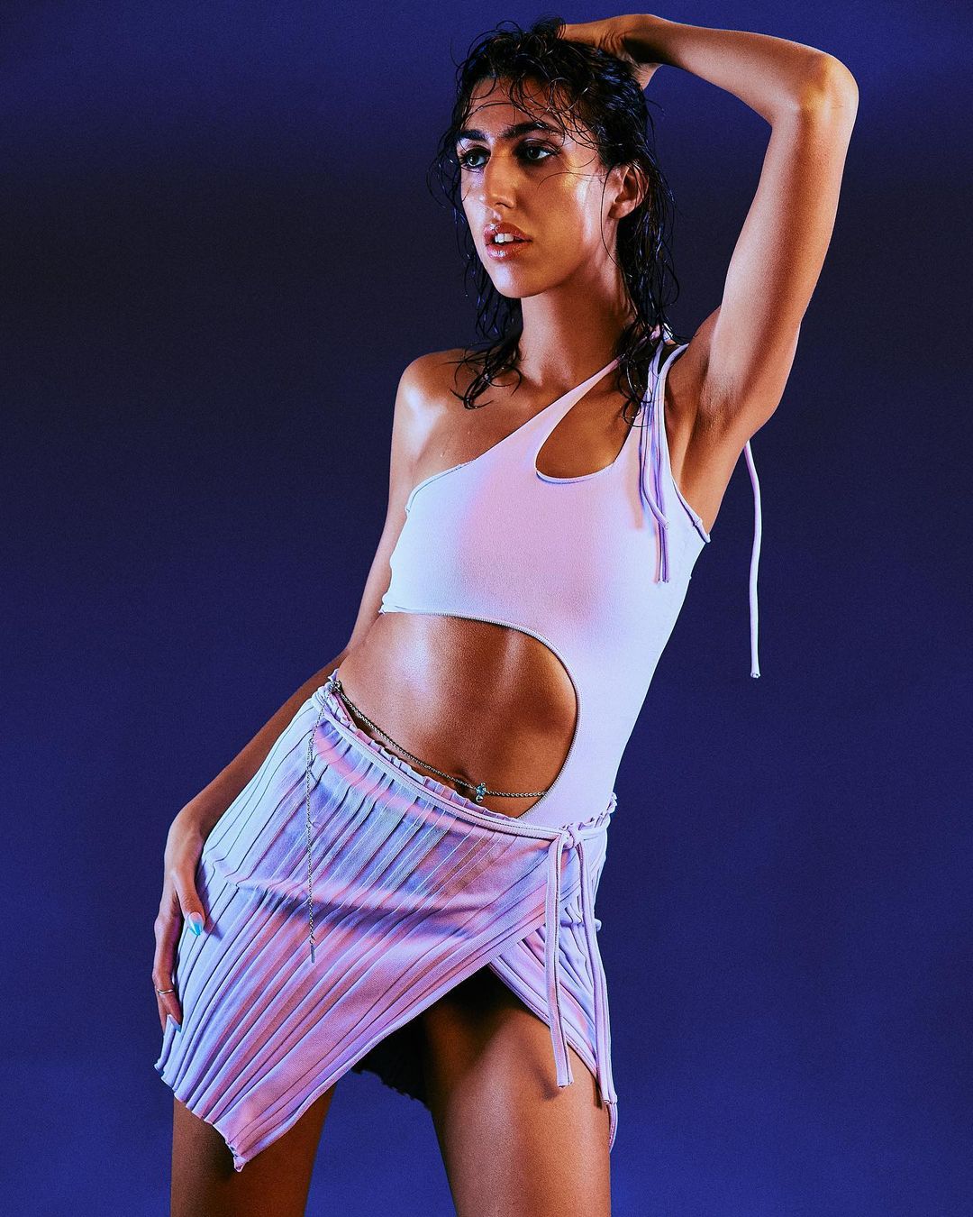 A woman poses in a one-shoulder top and pleated skirt against a purple background, fashionably lit in a studio environment.