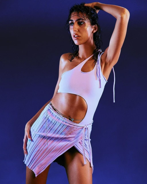 A woman poses in a one-shoulder top and pleated skirt against a purple background, fashionably lit in a studio environment.