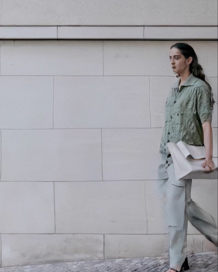 A woman in a green shirt and pants walks past a grey brick wall, carrying a large white clutch. A fashionable outdoor fashion shot.