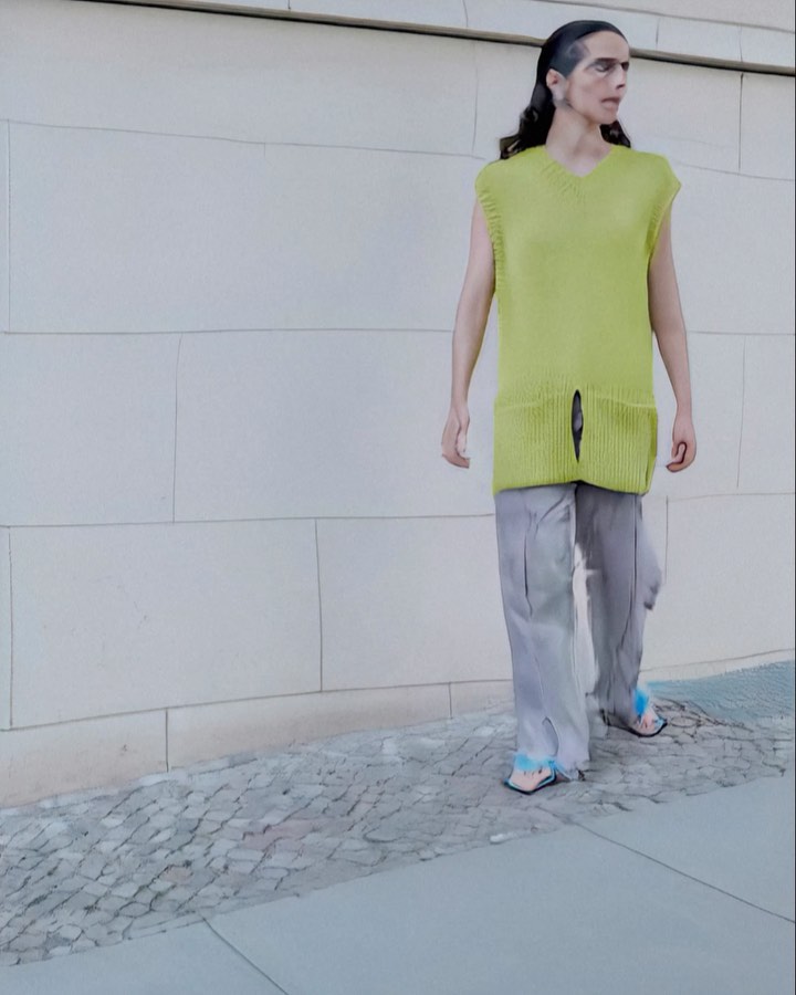 A person walks outside, wearing a chartreuse knit top and grey pants, on a stone sidewalk next to a textured, off-white wall.