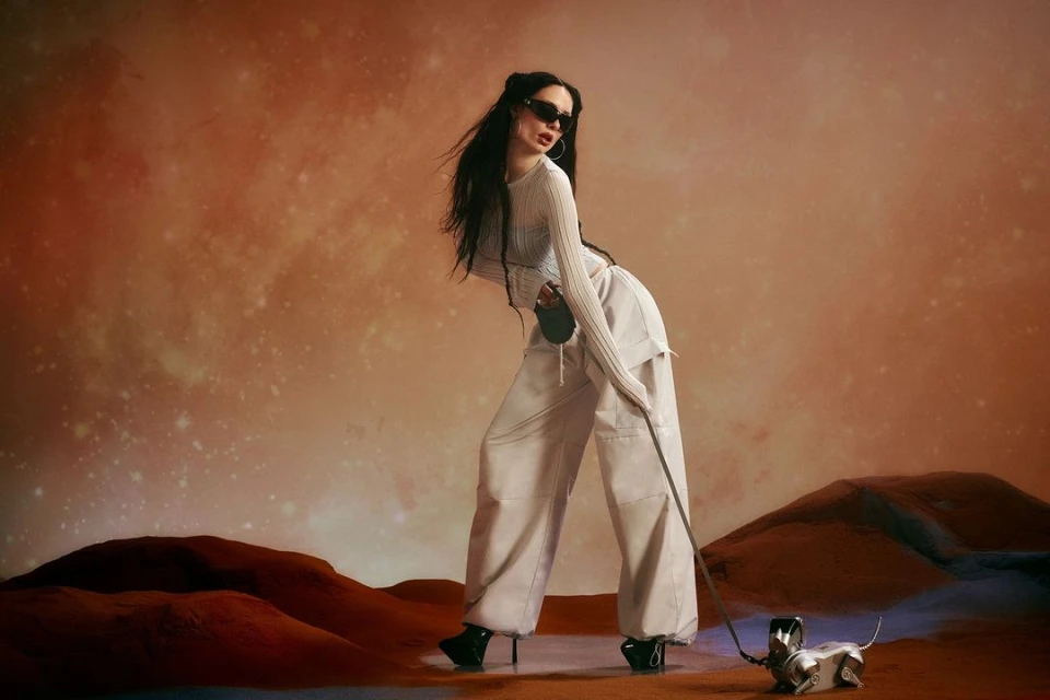 An adult woman poses with a metallic toy dog against a surreal desert backdrop in a fashion editorial photograph, showcasing modern style.