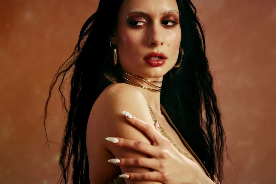 Portrait of a young adult woman with long, dark hair, wearing gold jewellery and makeup, posing against a mottled, brownish background.