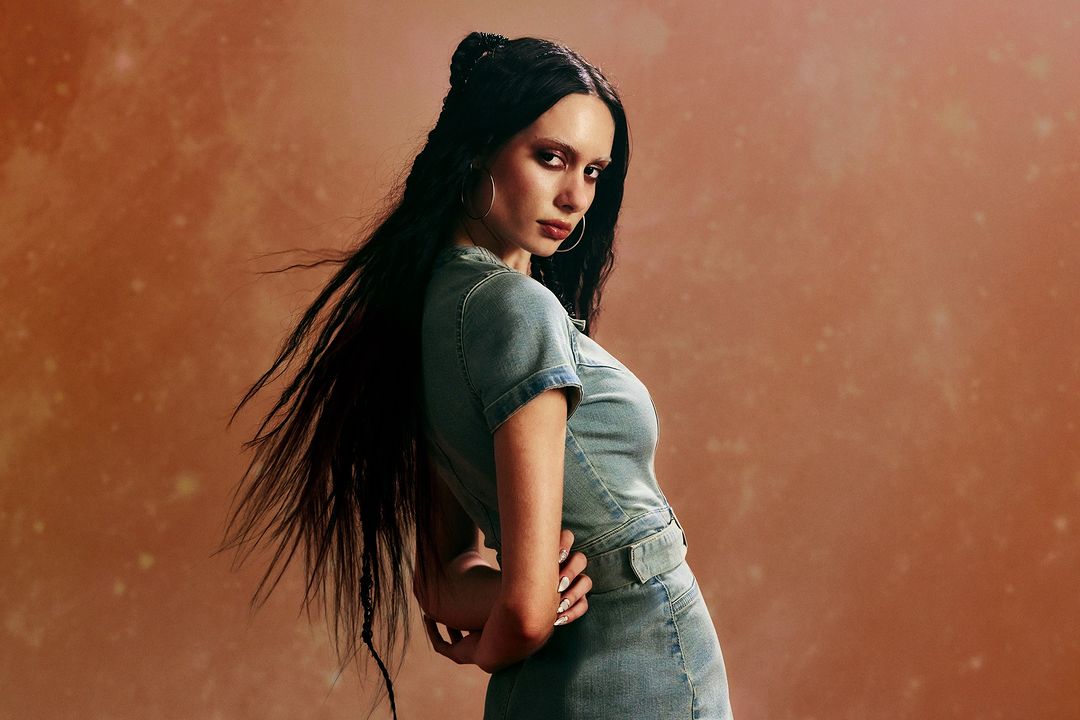 Portrait of a woman with long dark hair wearing a light denim mini dress and hoop earrings, posed against a mottled orange background.