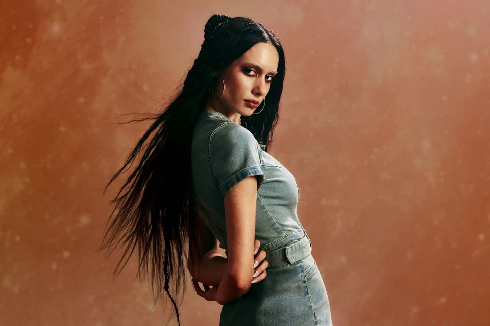 Portrait of a woman with long dark hair wearing a light denim mini dress and hoop earrings, posed against a mottled orange background.