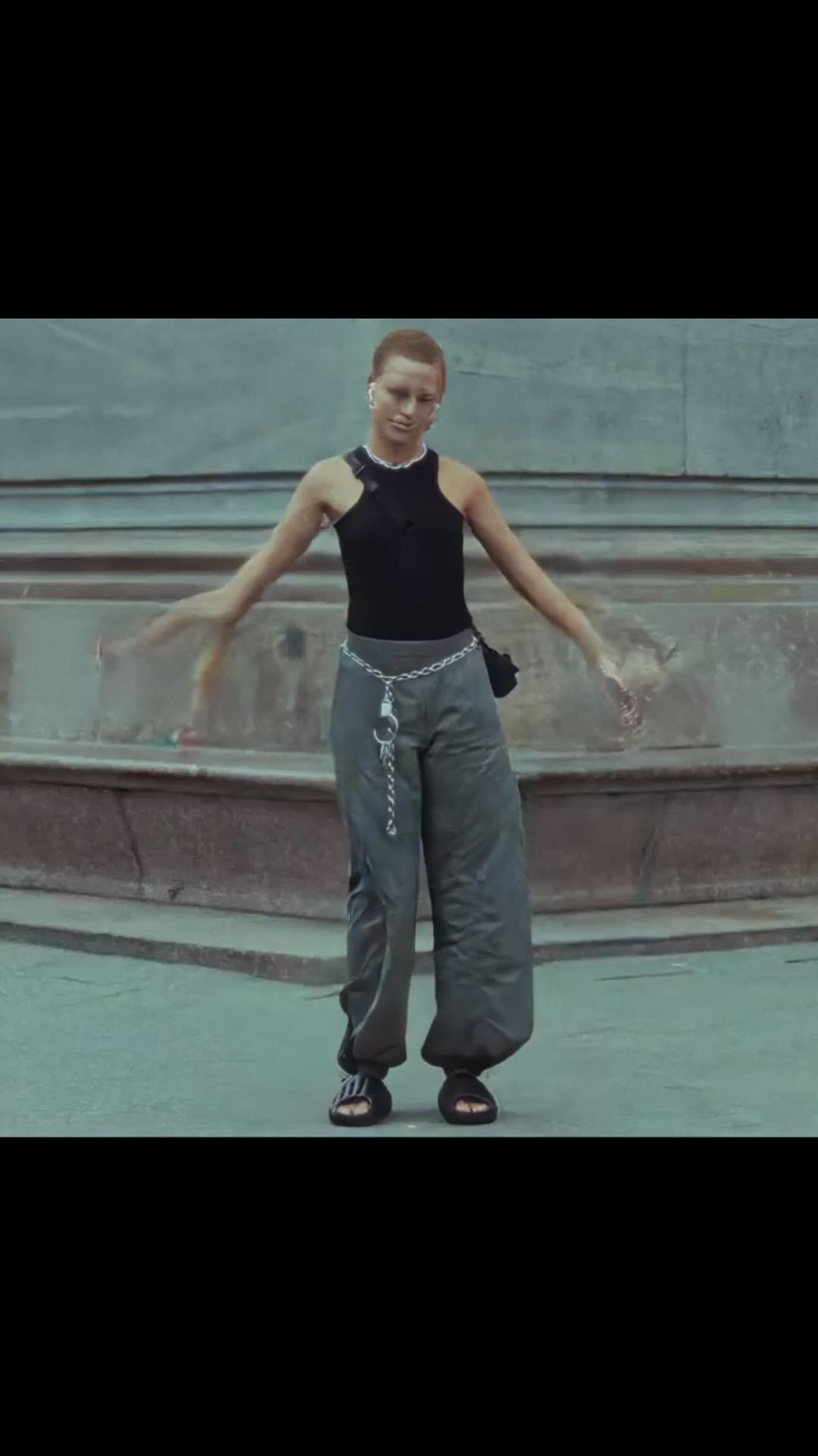 Young adult woman in tank top and parachute pants stands in a city square with her arms out, wearing a crossbody bag and platform sandals.
