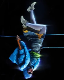 Model suspended upside down wearing a blue puffer jacket and sneakers against a black background with glowing light streaks.