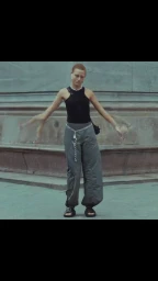 Model with short hair wearing a black tank top and grey trousers in front of a stone building exterior