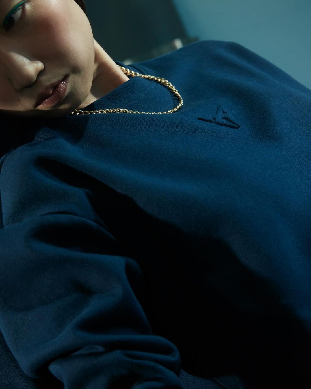 A close-up of a person with light skin and gold chain wearing a dark blue sweatshirt in a softly lit, neutral indoor setting.