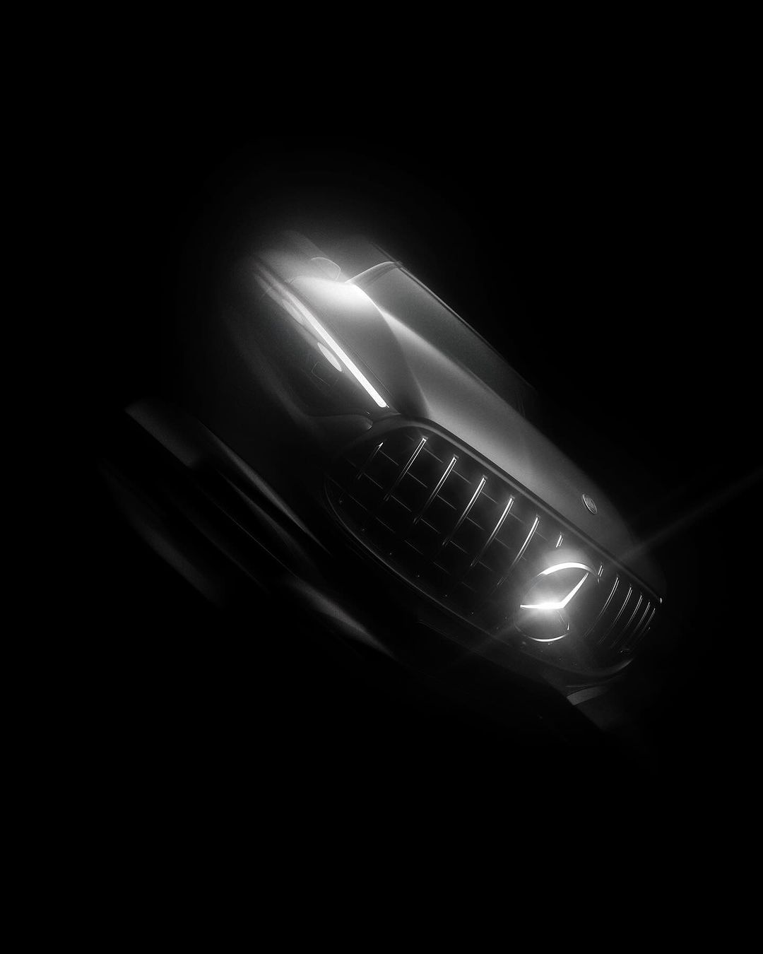 Close-up of the front grill and headlight of a silver car shown in dramatic lighting against a stark black background.