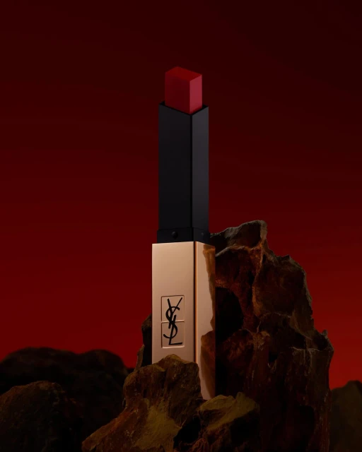 Close-up studio shot of YSL lipstick with a red hue, gold case, and the brand's logo, displayed on dark rocks against a blurred red background.