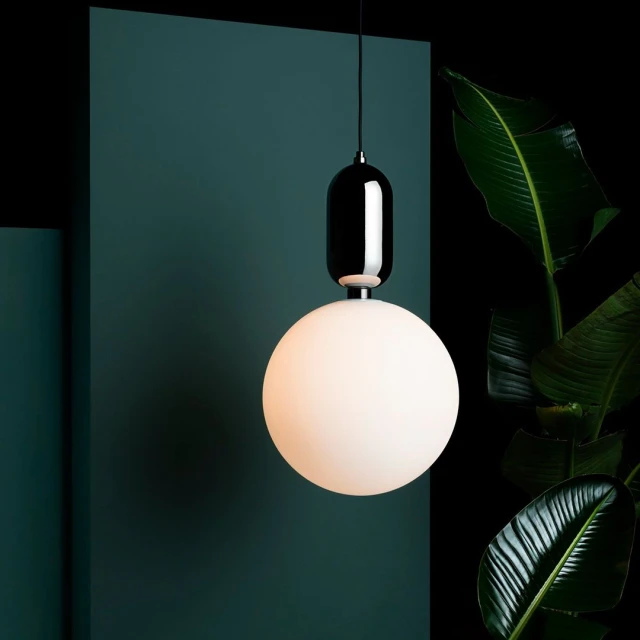 Modern black and white pendant light fixture with a spherical frosted glass shade, hanging against a dark green wall, with plant leaves.