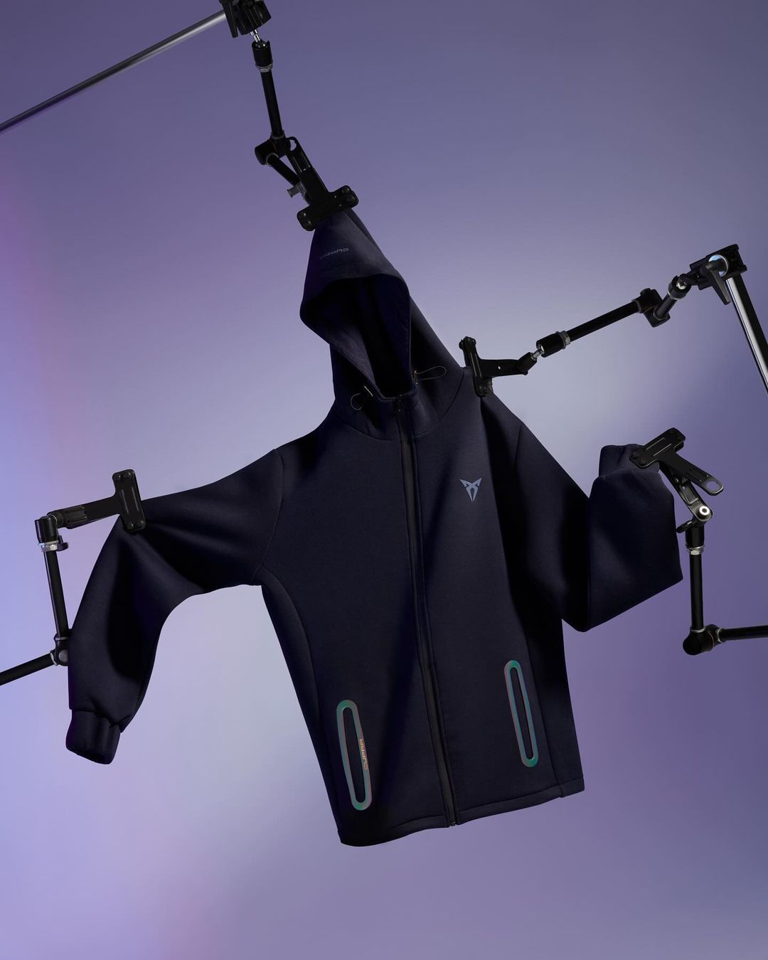 A dark blue hooded sweatshirt is displayed against a soft purple background, suspended by black articulating arms and clips.