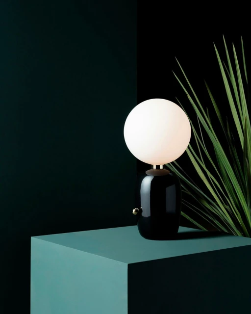 Indoor studio shot of a black table lamp with a white globe light on a teal block, set against a dark green and black background.