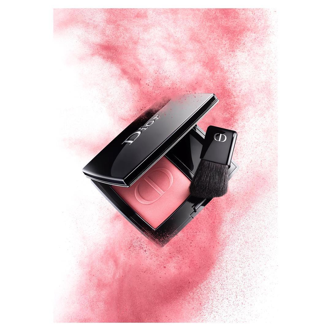 Dior blush palette with brush, showcasing the brand's logo and a pink powder, set against a soft pink powder cloud.