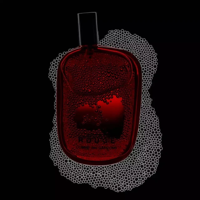 High-contrast still life of a red perfume bottle by Comme des Garçons surrounded by foam bubbles against a stark black background.
