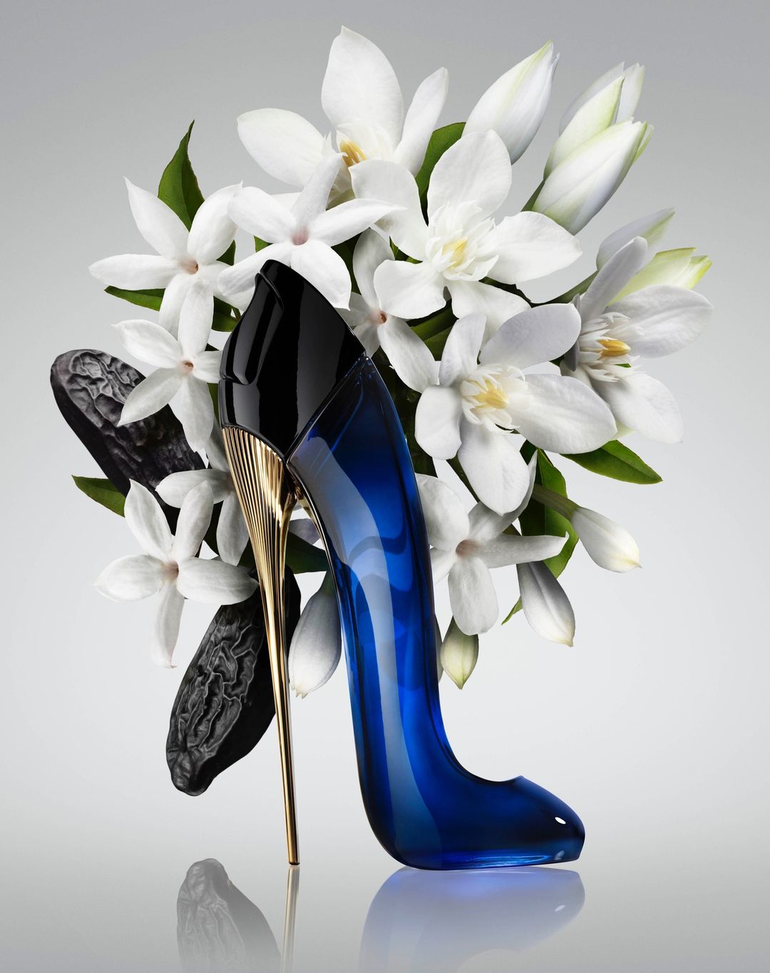 Elegant perfume bottle shaped like a stiletto heel, surrounded by white flowers and dark brown seed pods against a neutral background.