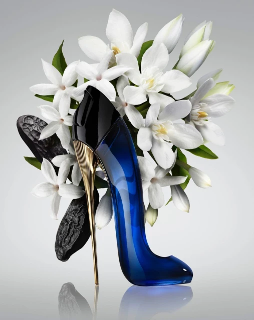 Elegant perfume bottle shaped like a stiletto heel, surrounded by white flowers and dark brown seed pods against a neutral background.