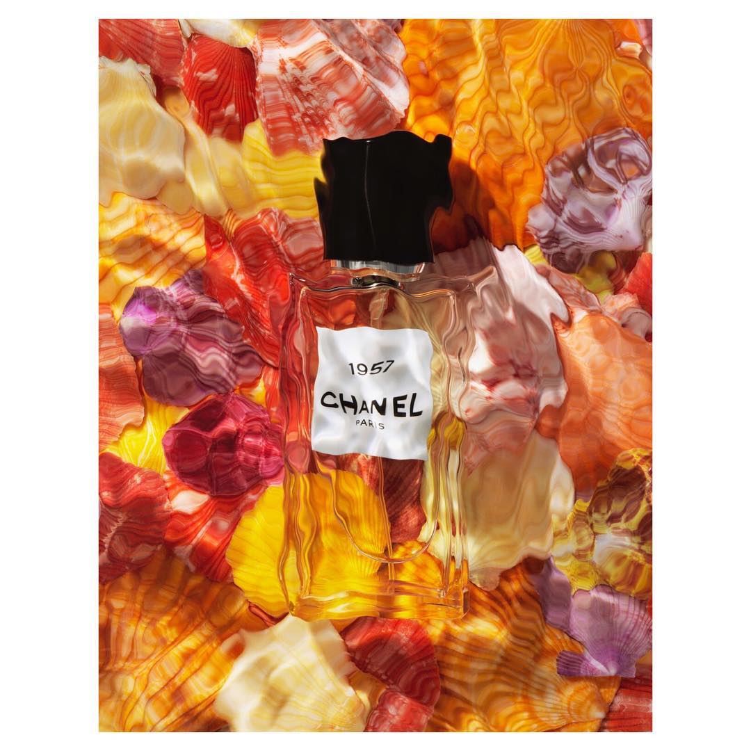 Chanel 1957 perfume bottle with black cap and white label sits in a bed of colorful seashell fragments with warm, diffused light.