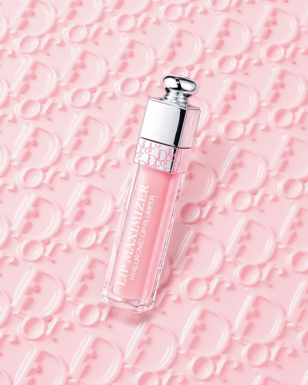 Dior Lip Maximizer on a pink background with embossed Dior logos. The lip gloss has a silver cap and pink tint.