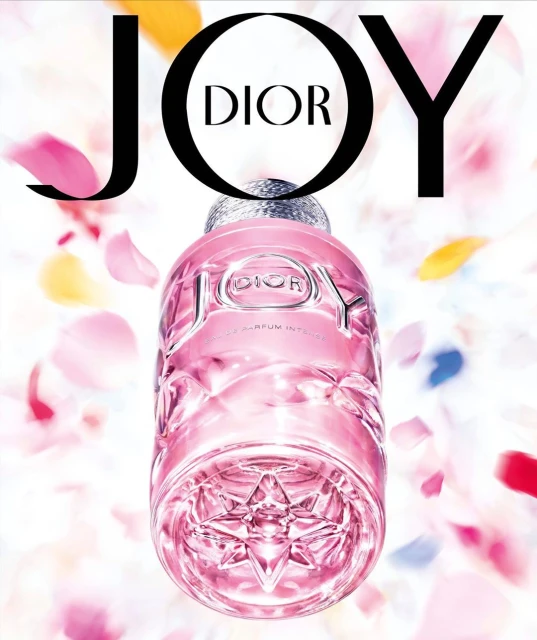 Close-up shot of a JOY by Dior perfume bottle with pink liquid against a soft-focus background of pink and yellow flower petals.