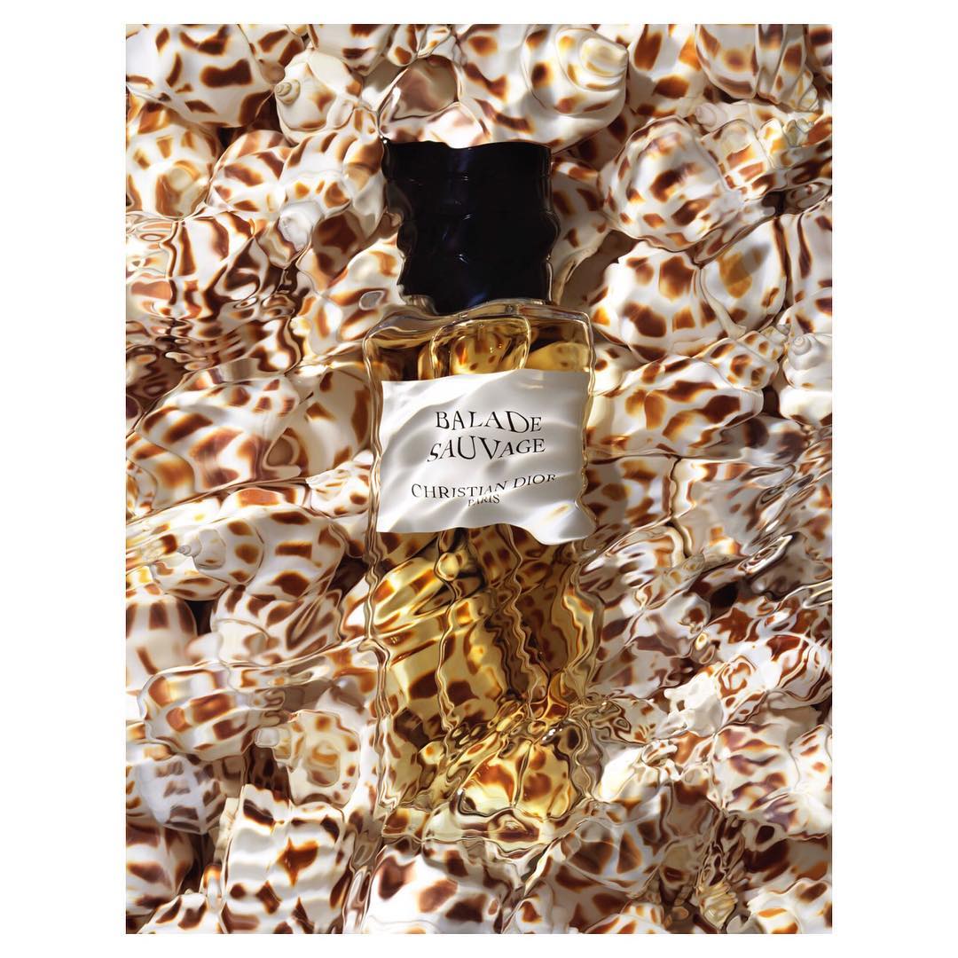 High-angle shot of a Balade Sauvage perfume bottle among a sea of shells, the light refracting through a shallow layer of water.