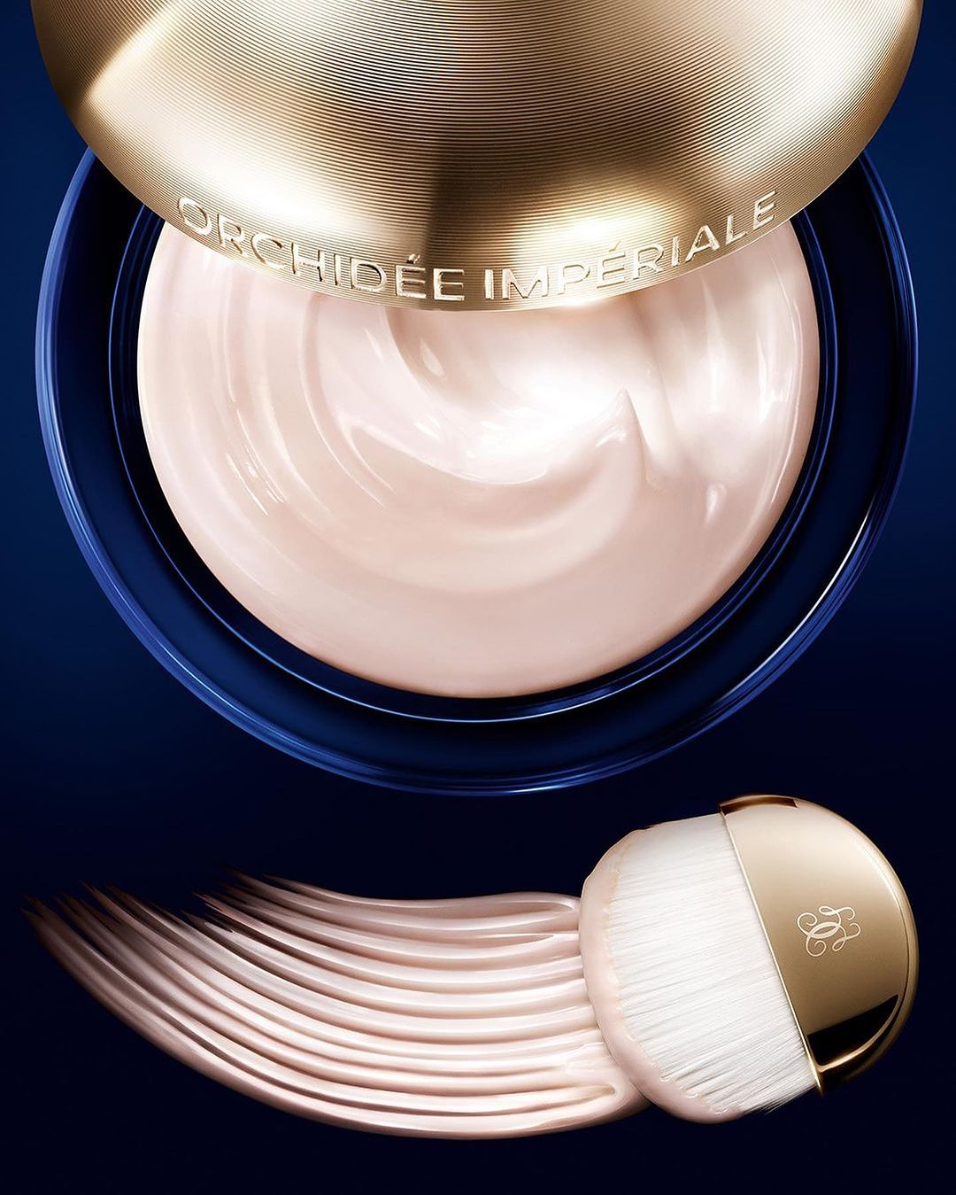 Close-up of Guerlain Orchidee Imperiale cream in a blue jar with gold lid and makeup brush on a dark blue background.