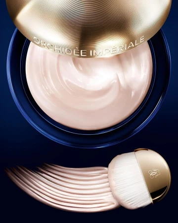 Close-up of Guerlain Orchidee Imperiale cream in a blue jar with gold lid and makeup brush on a dark blue background.