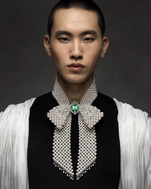Close-up fashion shot of an Asian male model with a shaved head wearing a black top and large pearl-encrusted bow-tie necklace with a green jewel.